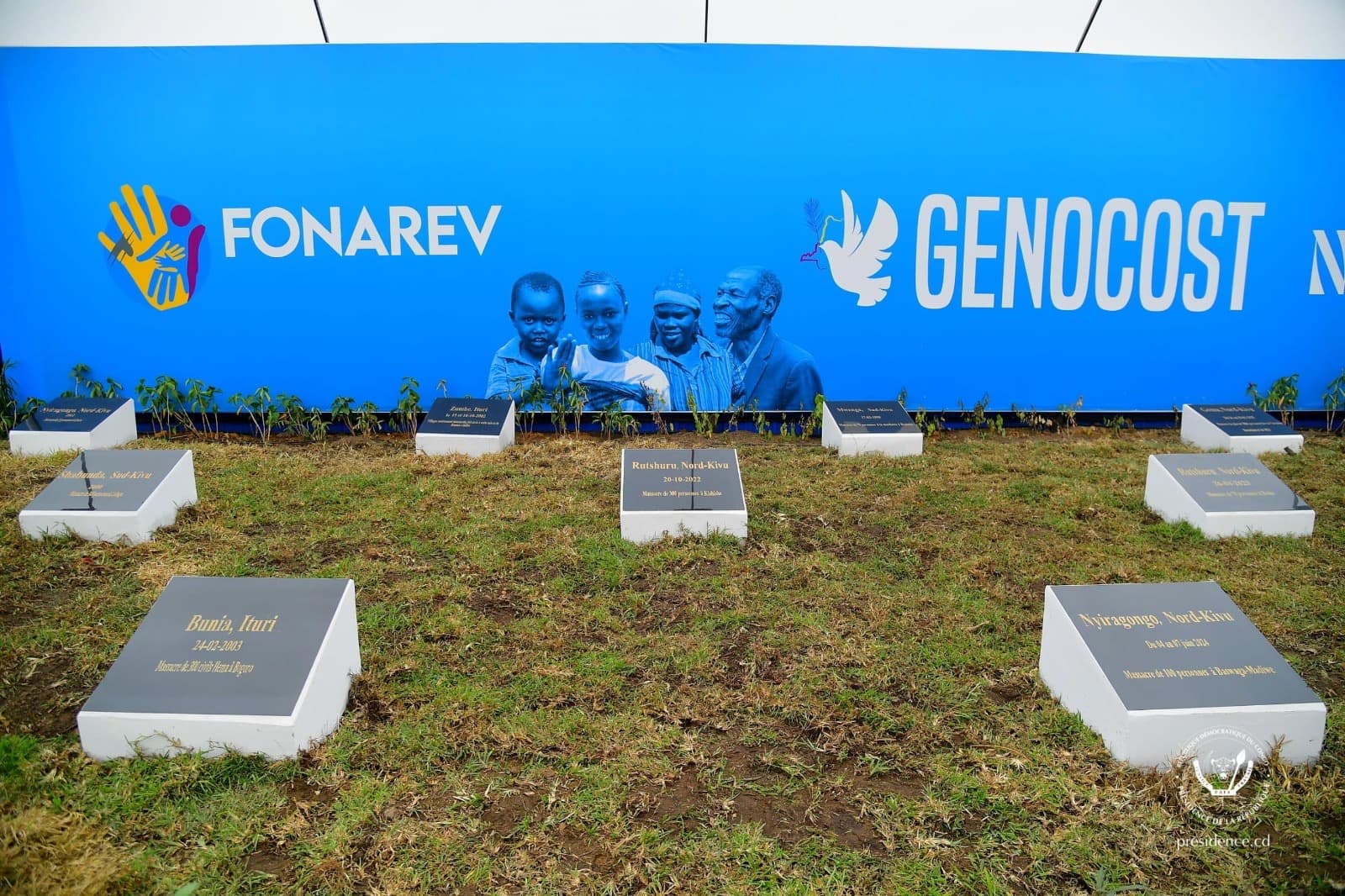 Installation FONAREV · GENOCOST — le néologisme apposé sur une fresque officielle avec des marqueurs funéraires symboliques